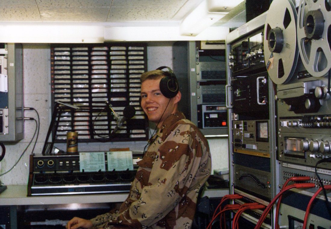 Adam Honoré in desert camouflage and headphones, smiling from the mixing console of a radio studio lined with tape cartridges and reel-to-reel decks.